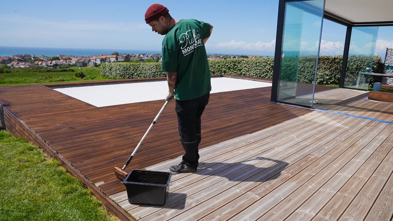 Person applying wood oil to a wooden deck with a brush and container, overlooking a scenic view.