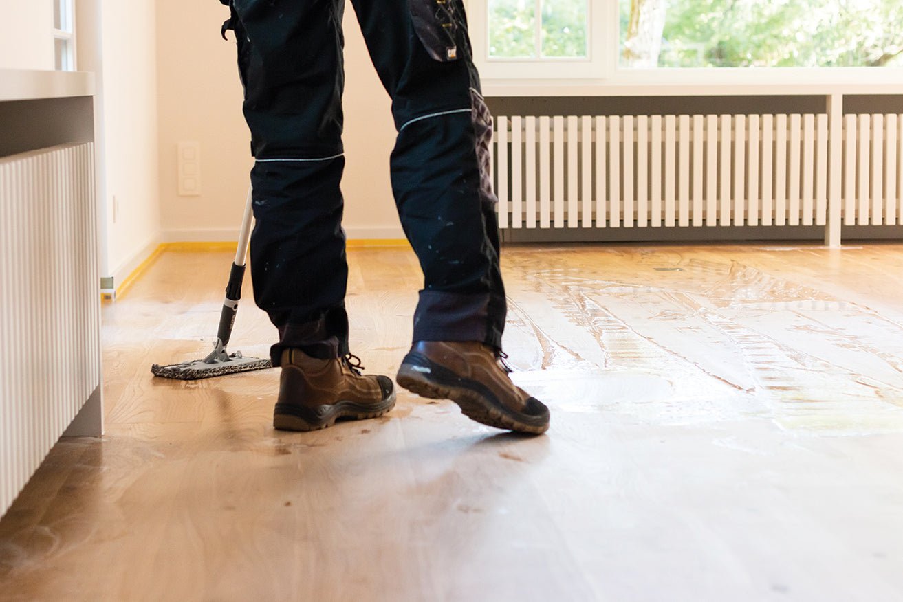 Person cleaning a wooden floor with a mop in a room with large windows.